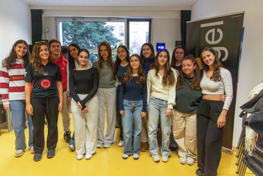 Gruppe von elf jungen Frauen steht in einem Raum mit gelbem Boden vor einem Fenster und einem Banner mit dem Schriftzug 'igel'.