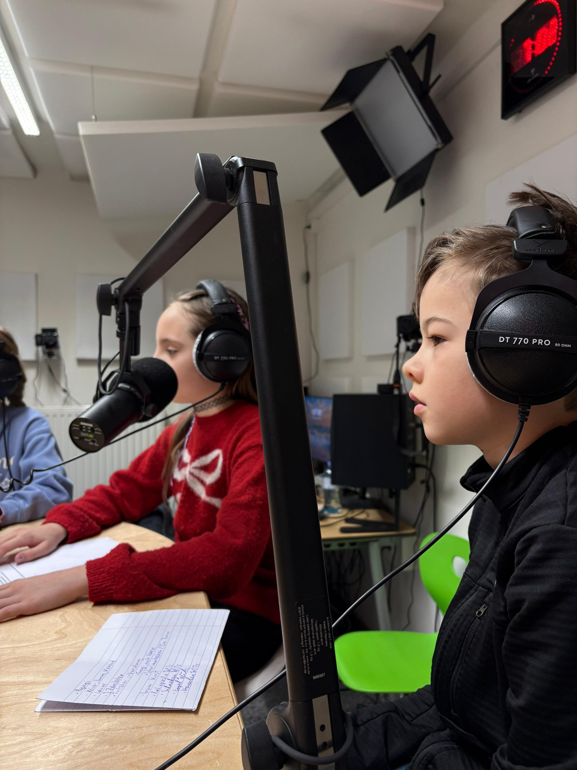 Zwei Personen mit Kopfhörern sitzen an einem Tisch mit Mikrofonen und Notizen in einem Tonstudio.