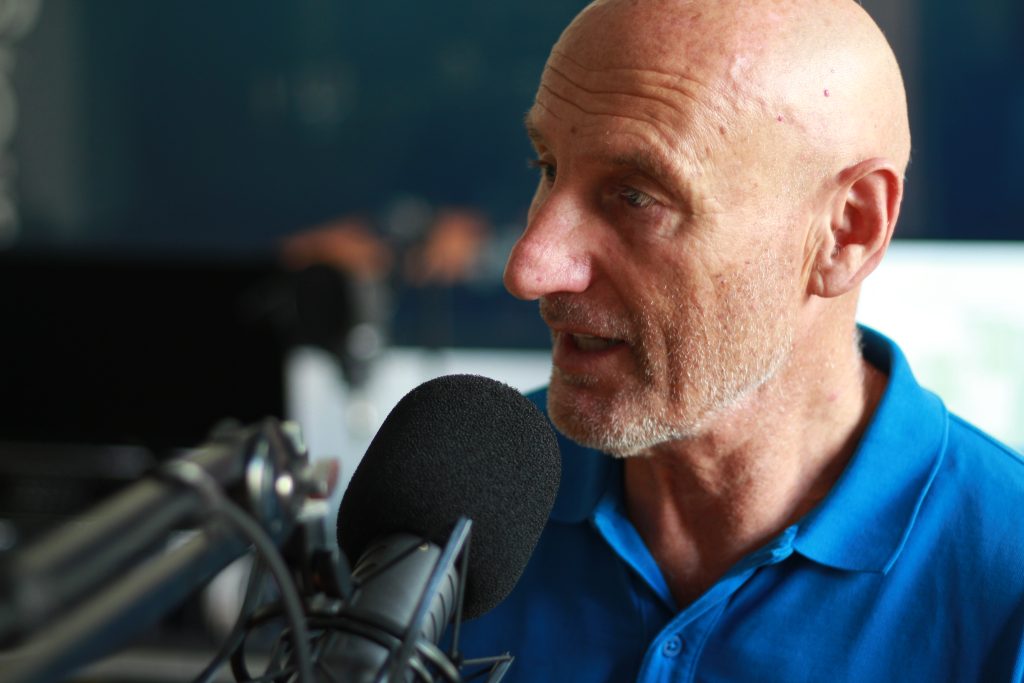 Mann mit blauem Poloshirt spricht in ein Mikrofon vor unscharfem Hintergrund mit technischer Ausrüstung.