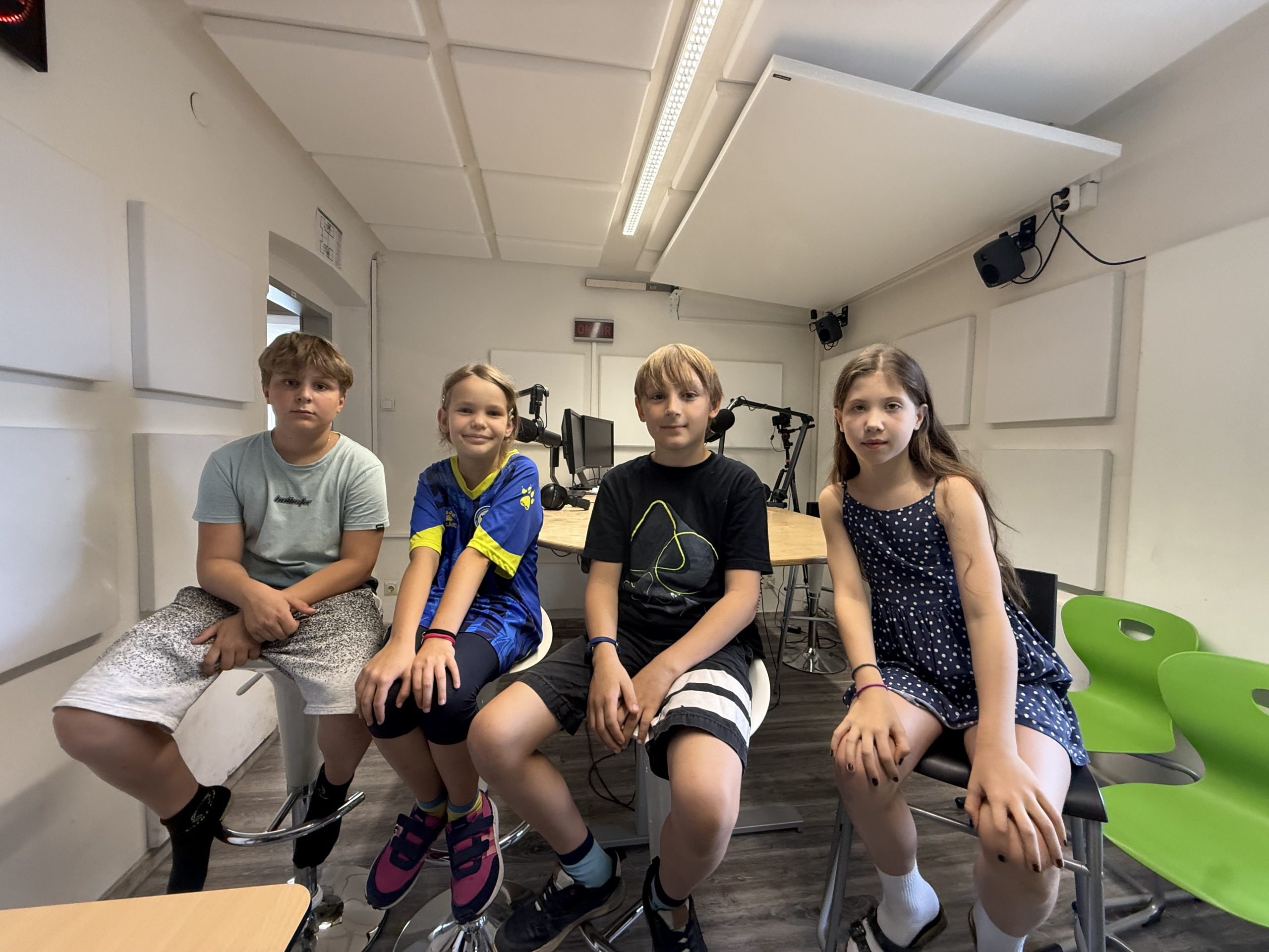 Vier Kinder sitzen nebeneinander auf Stühlen in einem Raum mit Mikrofonen und Akustikplatten an den Wänden