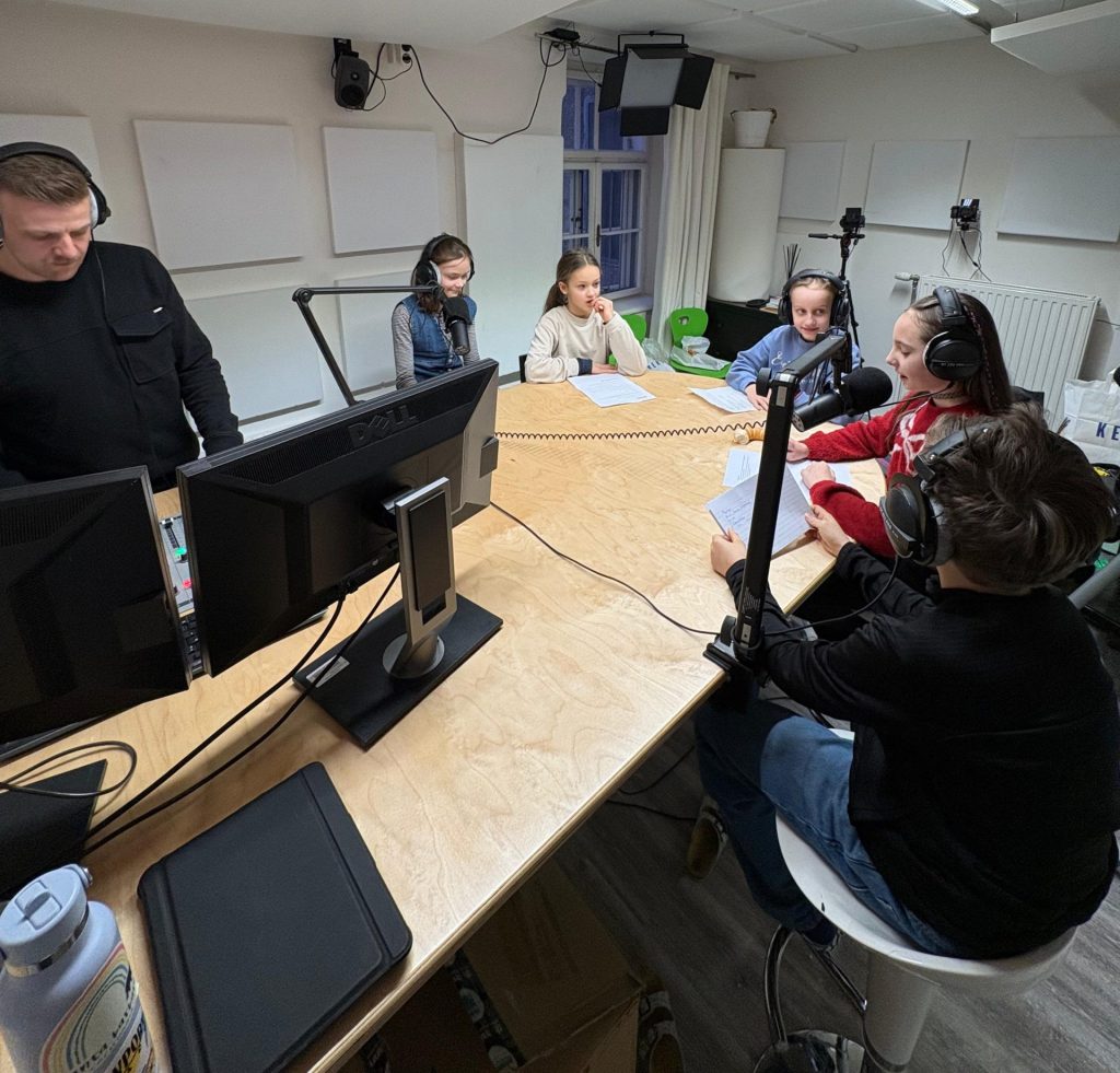 Mehrere Personen sitzen an einem großen Tisch in einem Radiostudio mit Mikrofonen, Kopfhörern und Computermonitoren.