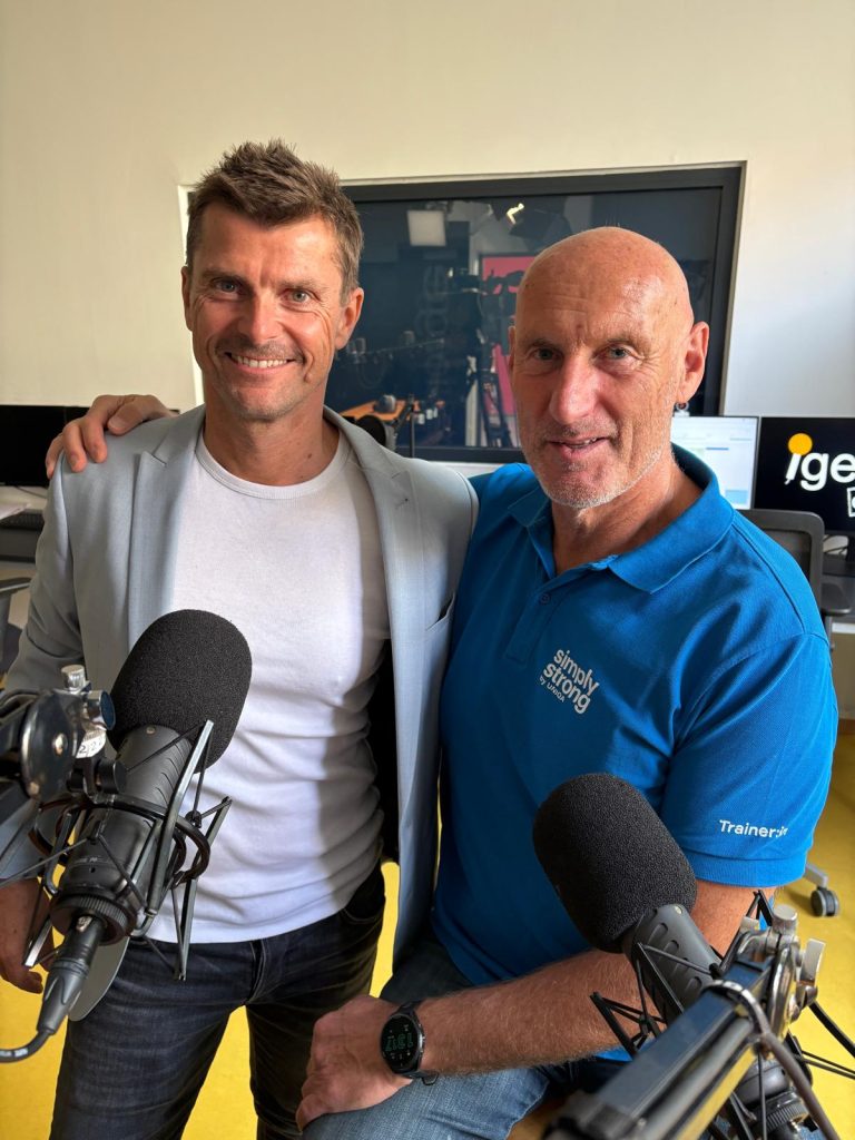 Zwei Männer stehen nebeneinander vor Mikrofonen, einer trägt ein graues Sakko und weißes T-Shirt, der andere ein blaues Polo mit Aufschrift 'simply strong' und 'Trainer'.