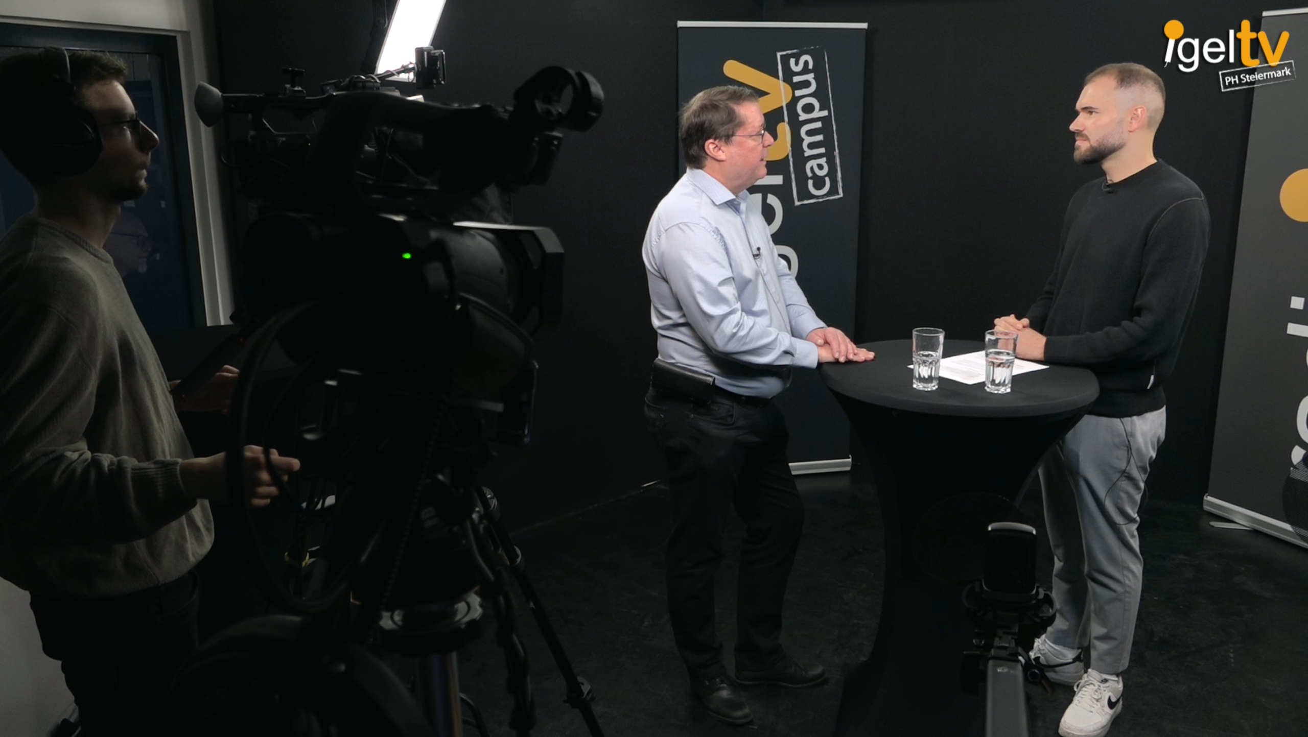 Zwei Männer stehen an einem Stehtisch mit Gläsern und sprechen in einem Studio, links eine Kamera und eine Person mit Kopfhörer.