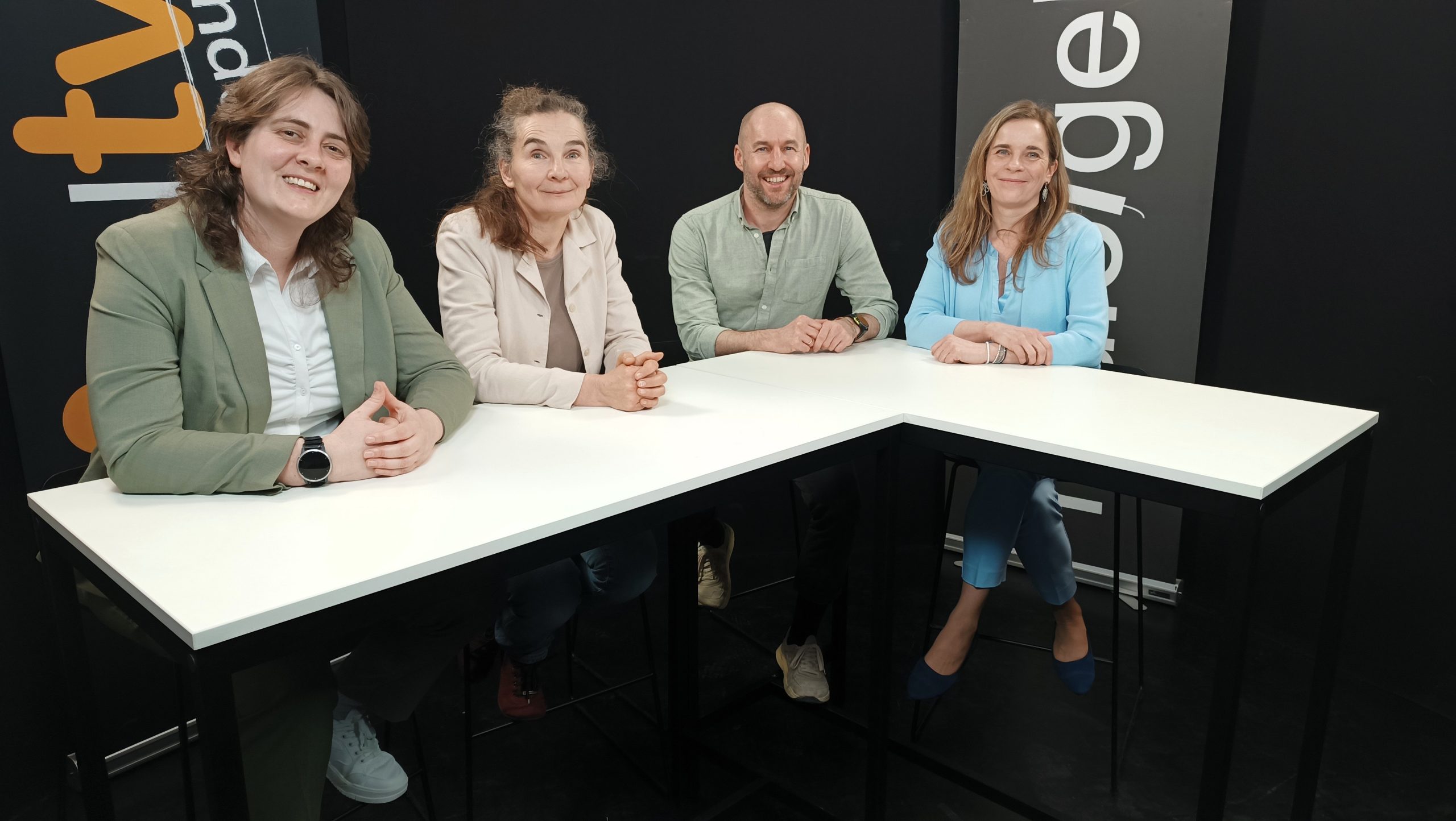 Vier Personen sitzen an einem weißen, eckigen Tisch vor schwarzem Hintergrund mit orange-weißer Schrift